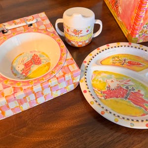 May include: A children's dinnerware set featuring a white bowl, a three-sectioned plate, a two-handled sippy cup, a fork, and a spoon. The items are decorated with colorful illustrations of rabbits, carrots, and checkered patterns in pink and yellow.