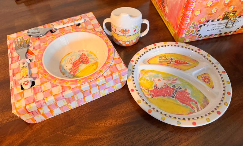 May include: A children's dinnerware set featuring a white bowl, a three-sectioned plate, a two-handled sippy cup, a fork, and a spoon. The items are decorated with colorful illustrations of rabbits, carrots, and checkered patterns in pink and yellow.