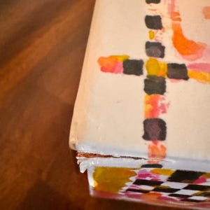 May include: A rectangular box with a white surface featuring a hand-painted design of black, orange, and pink squares arranged in a cross pattern. The edges of the box show signs of wear with slightly peeling paper layers at the corner seam.