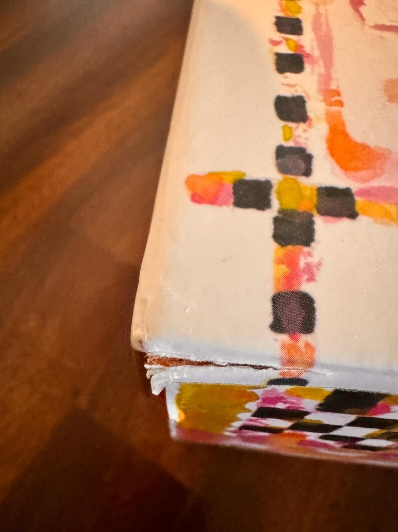 May include: A rectangular box with a white surface featuring a hand-painted design of black, orange, and pink squares arranged in a cross pattern. The edges of the box show signs of wear with slightly peeling paper layers at the corner seam.