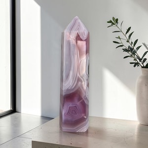 Puede incluir: Una torre de cristal pulido alta que presenta capas translúcidas de bandas de color rosa claro, lavanda y blanco. La piedra tiene una punta hexagonal en la parte superior y exhibe patrones de ágata naturales y arremolinados en su estructura.