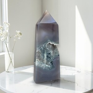 Puede incluir: Un alto obelisco de cristal pulido gris-púrpura con una cavidad de geoda. El cristal se exhibe sobre una mesa de mármol blanco. La cavidad de la geoda revela un grupo de cristales azul-gris. Un jarrón de flores blancas está en el fondo.