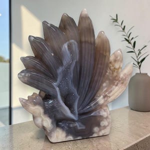 Op de afbeelding: Een sculptuur van gepolijst agaat met een waaiervormige opstelling van gelaagde, doorschijnende grijze en beige bloemblaadjes. Het midden van het stuk onthult een diepe, getextureerde geodeholte gevuld met kleine, sprankelende kristallen.