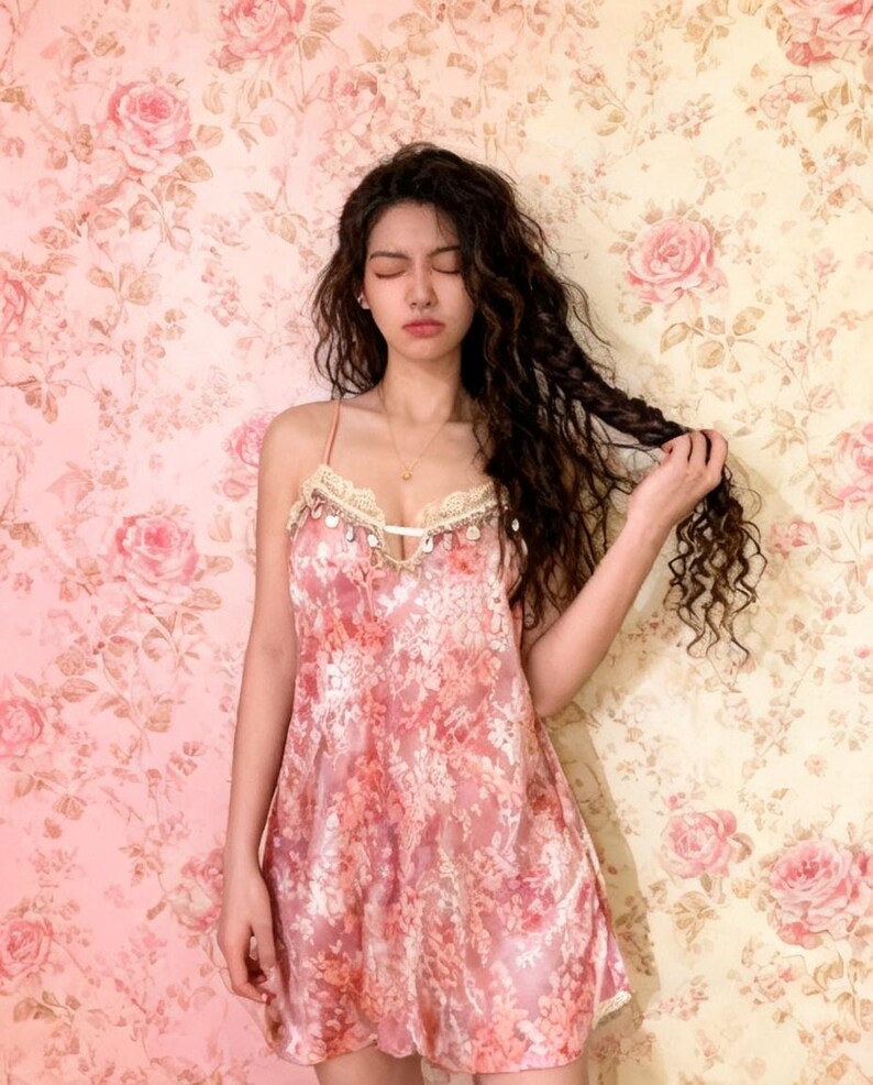 May include: A pink floral print slip dress with spaghetti straps and lace trim. The dress has a V-neckline and a loose, flowing silhouette. The background is a floral wallpaper in shades of pink and cream.