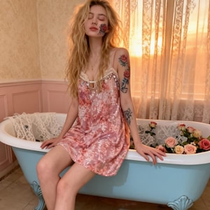 May include: A pink floral print slip dress with spaghetti straps. The dress has a delicate lace trim and is knee-length. The model is sitting on the edge of a light blue clawfoot bathtub filled with roses. The background features a window with sheer curtains.