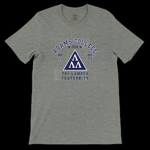 Peut inclure: Un t-shirt gris avec le texte bleu marine "ADAMS COLLEGE USA" dans un design en arc. En dessous, un triangle bleu marine avec les lettres grecques "ΛΛ". Le texte "TRI-LAMBDA FRATERNITY" est en dessous.