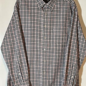 May include: A long-sleeved button-down shirt with a checkered pattern in black, white, red, and green. The shirt has a classic collar, buttoned cuffs, and a front pocket. The shirt is hanging on a black hanger.