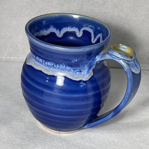 Op de afbeelding: Een ronde, keramische mok met een diepblauwe glazuurlaag en een horizontale ribbeltextuur. De rand heeft een wit, druipend glaze-effect dat langs de binnen- en buitenkant loopt. Een stevig handvat is aan de zijkant bevestigd met een duimsteun.