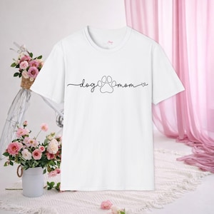 Puede incluir: Camiseta blanca con las palabras "dog mom" en fuente cursiva, una huella de pata y un corazón. El fondo incluye una bicicleta con una cesta de flores rosas y una cortina rosa.
