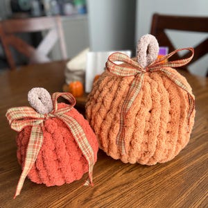 May include: Two handmade plush pumpkins crafted from chunky orange and light orange yarn. Each pumpkin features a soft, light brown yarn stem and is decorated with a tied plaid ribbon in shades of orange, green, and brown around the top.