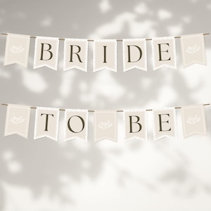 Pode incluir: Duas faixas com as palavras "BRIDE" e "TO BE" impressas em castanho escuro em bandeirolas creme. Cada bandeirola tem um desenho de coroa decorativa. As faixas são presas em cordas finas castanhas.
