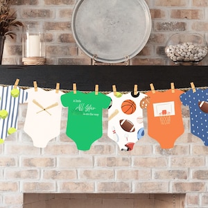 Op de afbeelding: Een decoratieve slinger met zes papieren uitsparingen in de vorm van babyrompertjes, elk met een uniek sportthema, waaronder tennisballen, honkbalknuppels, een groen rompertje met de tekst A little All Star is on the way, basketballen en voetballen.