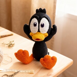 May include: A handmade black and orange crochet duck doll with large white eyes and a yellow beak. The duck has outstretched arms and orange feet. The doll is sitting on a light wooden surface, with crafting supplies in the background.