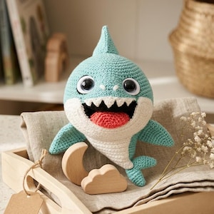 May include: A crocheted shark plush toy with a light blue body, a white belly, large black eyes, and a wide open mouth showing white teeth and a red tongue. The shark is positioned next to small wooden moon and cloud shapes in a wooden tray.