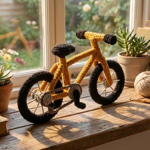 May include: A handmade yellow crochet bicycle with black wheels and a seat, set on a wooden windowsill. The miniature bike is next to potted plants, books, and a ball of yarn, creating a decorative scene.