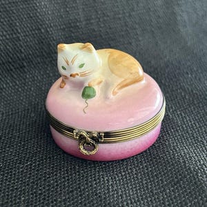 May include: A round porcelain trinket box featuring a white and orange cat resting on a pink lid. The cat is playing with a small green ball of yarn. The box has a brass-colored metal hinge with a decorative ring-shaped clasp on the front.