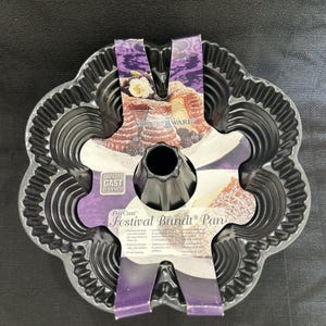 Op de afbeelding: Een donkergrijze Festival Bundt-pan van zwaar gegoten aluminium met een gegroefd, bloemachtig ontwerp. De pan heeft een centrale buis en is verpakt met een paars en wit label van Nordic Ware.