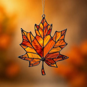 May include: A stained glass maple leaf ornament featuring variegated shades of orange, yellow, and deep red glass segments held together by dark metal solder lines. The leaf hangs from a thin string attached to a small metal ring at the top.