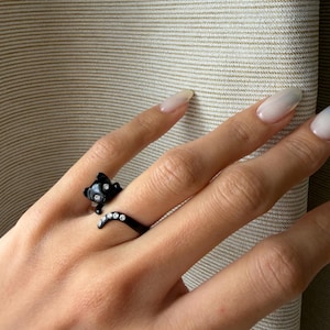 May include: A black cat ring with a detailed design. The cat's head is at the top of the ring, and its tail curves around the finger. The ring features small, sparkling accents.