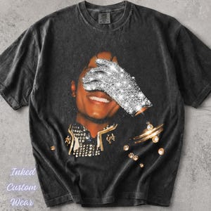 Puede incluir: Camiseta gris oscuro con un retrato de una persona, un ojo cubierto por un guante brillante. La camiseta tiene adornos dorados y el texto "Inked Custom Wear" en una fuente cursiva.