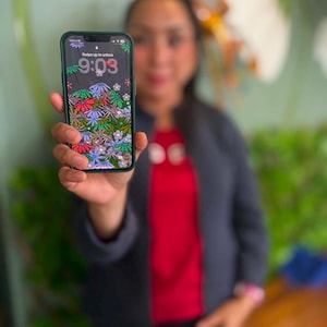 May include: A smartphone with a dark green case displaying a lock screen with a colorful floral pattern, the time 9:03, and the text Swipe up to unlock. The screen features a dense arrangement of stylized flowers in shades of red, blue, and white.