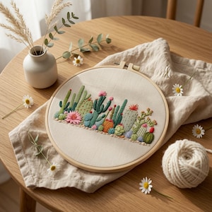 Puede incluir: Un bastidor de bordado circular de madera que contiene una tela de color blanco roto con un diseño detallado bordado a mano de varios cactus y suculentas verdes, con detalles florales en rosa, naranja y rojo en una fila horizontal.