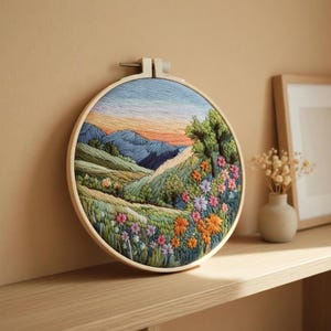 May include: A round embroidery hoop featuring a colorful landscape scene. The artwork depicts rolling green hills, blue mountains, and a vibrant array of flowers in shades of orange, pink, and purple. The sky transitions from blue to orange.