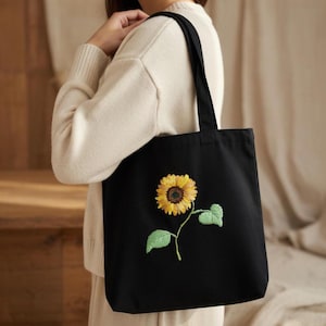 Pode incluir: Bolsa tote preta com um girassol bordado. O girassol tem pétalas amarelas, um centro castanho e folhas e caule verdes. A bolsa tem duas alças pretas.