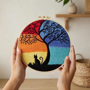 May include: A colorful embroidered hoop art featuring a tree and a cat silhouette. The design is divided into four sections with red, blue, orange, and yellow hues. The cat sits under the tree, which has black branches.