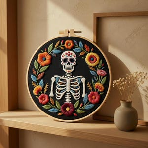 May include: Embroidered Day of the Dead skull and skeleton hoop art. The black fabric is surrounded by colorful flowers in orange, red, and yellow. The skeleton has a white skull with decorative details. The wooden hoop is visible.