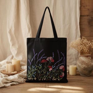 Op de afbeelding: Zwarte tote bag met geborduurd bloemendesign. De borduursels tonen lavendel, rode en roze bloemen met groene bladeren. De tas heeft twee zwarte handvatten en is gemaakt van een stevige stof.