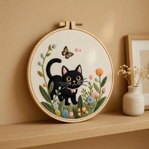 May include: A circular embroidery hoop displays a black cat with a pink collar, set amidst colourful flowers and a butterfly. The artwork is set against a cream-coloured background. The wooden hoop adds a rustic touch to the handcrafted piece.