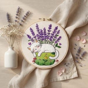 Puede incluir: Arte de aro bordado con un gato blanco durmiendo sobre un parche de hierba verde con lavanda púrpura. El gato tiene los ojos cerrados y las mejillas rosadas. El bordado está en un aro de madera, con un fondo de tela neutra.