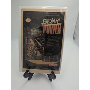 May include: A rectangular print featuring the text Bionic Power in large tan letters at the top. Below, a smaller image of the Bionic Commando video game box art by Capcom is displayed, showing a character in a combat scene. The background is black.