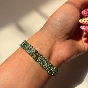 May include: A green rhinestone bracelet with three rows of sparkling stones. The bracelet is worn on a wrist, showcasing the jewelry's design and color. The bracelet's green stones catch the light, creating a shimmering effect.