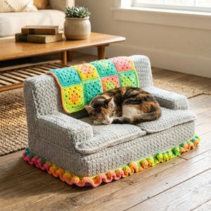May include: A gray crocheted cat sofa with a colorful blanket and a sleeping cat. The sofa has a rainbow trim along the base. The blanket is made of colorful squares. The cat is brown, black, and white.