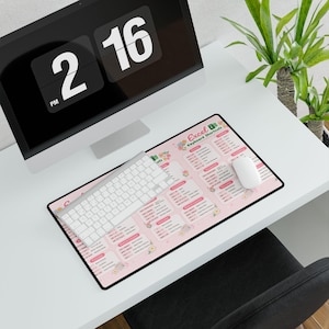 Può includere: Un tappetino per mouse rosa con bordo nero con testo "Excel Keyboard Shortcuts" e illustrazioni floreali. Una tastiera e un mouse bianchi sono sul tappetino, con un monitor di computer sullo sfondo. Il monitor mostra l'ora 14:16.