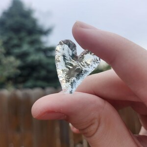 May include: A small, translucent, heart-shaped object made of clear glass or crystal. The surface has a textured, jagged appearance with light reflecting off the edges, creating a bright, shimmering effect against the natural outdoor light.