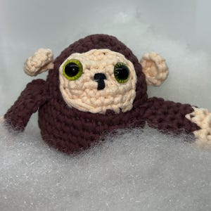 May include: A handmade crochet monkey with a brown body, beige face, and large green eyes. The monkey has small beige ears and arms. The toy is made of soft yarn and is designed to be a cuddly plushie.