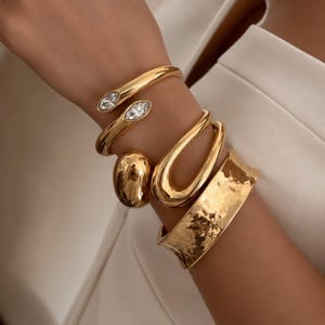 Könnte beinhalten: Eine Sammlung goldfarbener Armreifen, bestehend aus einem offenen Armreif mit klaren marquise-geschliffenen Steinen, einem glatten, kieselsteinförmigen Armreif, einem großen ovalen Armreif und einem breiten Armreif mit gehämmertem Metallfinish.