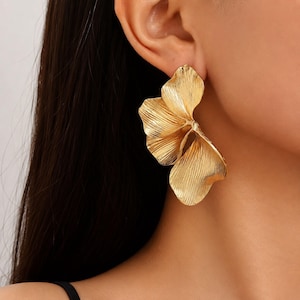Può includere: Un orecchino color oro caratterizzato da un grande design a fiore testurizzato con petali multipli. La superficie metallica presenta una finitura spazzolata e organica con sottili linee visibili che si irradiano dal centro, creando un effetto.