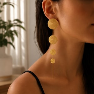 Peut inclure: Une longue boucle d'oreille pendante dorée composée de quatre disques circulaires de tailles variées disposés verticalement. Chaque disque présente une finition texturée et mate, reliés par une fine chaîne dorée délicate.