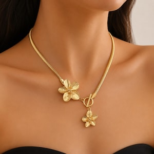 Può includere: Una collana color oro con una spessa catena a serpente, una chiusura a barra e due ciondoli a forma di fiore testurizzato. Un fiore è fissato alla catena vicino alla chiusura, mentre il secondo fiore pende dalla barra come un ciondolo.