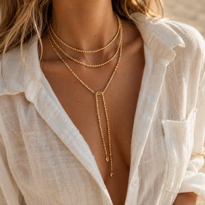 Puede incluir: Un collar de varias cadenas en tono dorado con pequeñas cuentas esféricas. El diseño incluye dos capas circulares superiores y una sección inferior estilo lazo que termina en dos pequeños dijes colgantes en forma de corazón.
