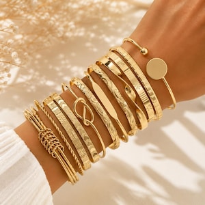 Op de afbeelding: Een set goudkleurige armbanden met diverse ontwerpen, waaronder een gladde ronde bedel, een gedraaide touwstructuur, een knoopdetail, een platte staaf en patronen met geometrische en florale motieven, allemaal samen gedragen als een gestapelde collectie.