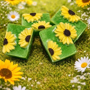 May include: Green rectangular soaps decorated with yellow sunflowers with black centres. The soaps are arranged on a bed of green grass with daisies and sunflowers in the background. The soaps are a decorative item.