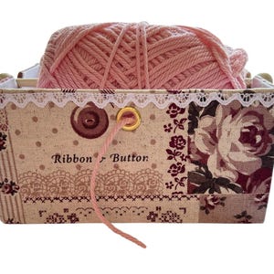 May include: A rectangular fabric basket with a floral and lace design, holding a ball of pink yarn. The basket has the words "Ribbon Button" printed on the front. The basket is ideal for storing craft supplies.