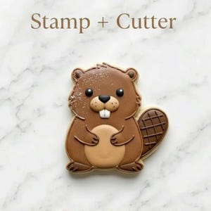 May include: A brown beaver-shaped cookie cutter and stamp set. The cookie is decorated with brown icing, a light brown belly, and a checkered tail. The text "Stamp + Cutter" is at the top.