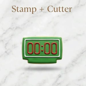 May include: Green cookie cutter and stamp in the shape of a digital clock displaying "00:00". The cookie has a white border and red numbers. The words "Stamp + Cutter" are above the cookie.