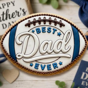 Puede incluir: Una galleta ovalada decorada con glaseado blanco, azul y marrón que imita un balón de fútbol americano. El centro presenta el texto BEST DAD EVER en letras azules y blancas. La galleta tiene un borde y cordones de color marrón.
